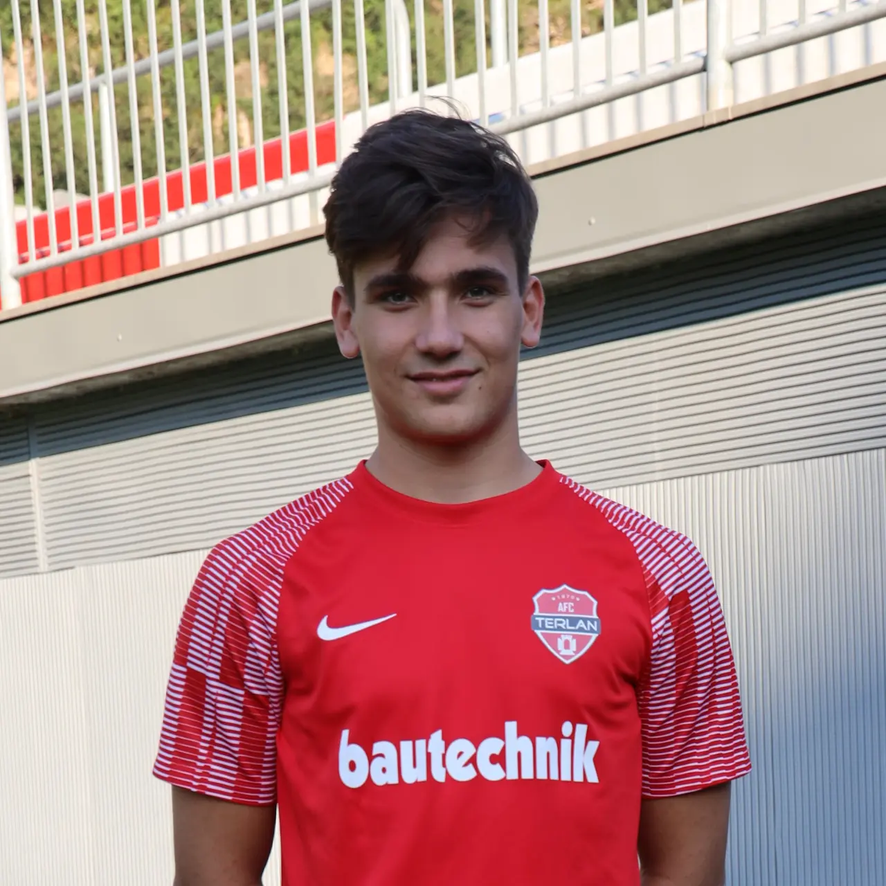 Porträt eines jungen Fußballspielers im roten AFC-Terlan-Trikot mit Bautechnik-Sponsor, vor einer grauen Wand und einem Geländer.