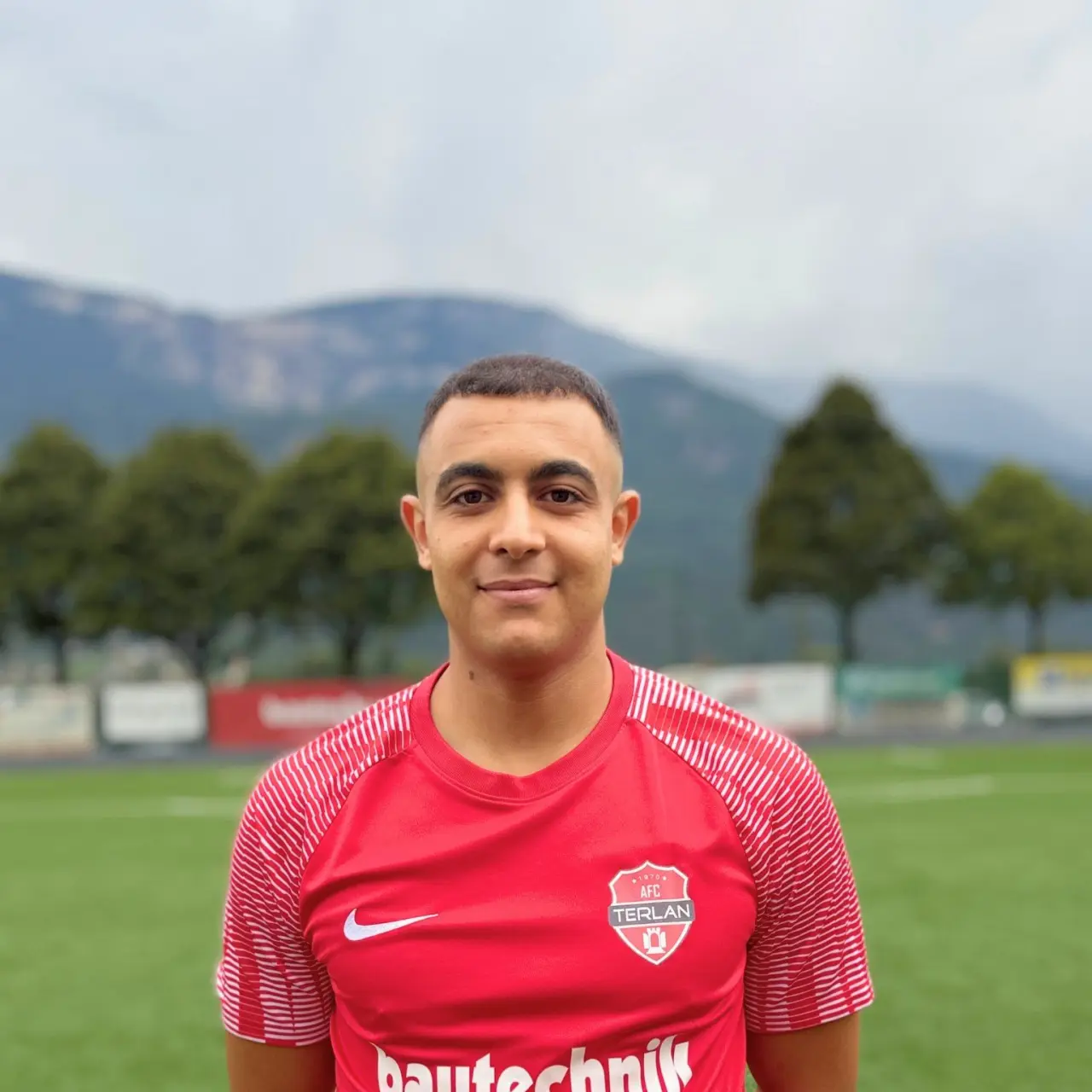 Porträt von Mahdi Sammah im roten AFC-Terlan-Trikot mit bautechnik-Sponsor auf einem Kunstrasenplatz, Berge im Hintergrund.