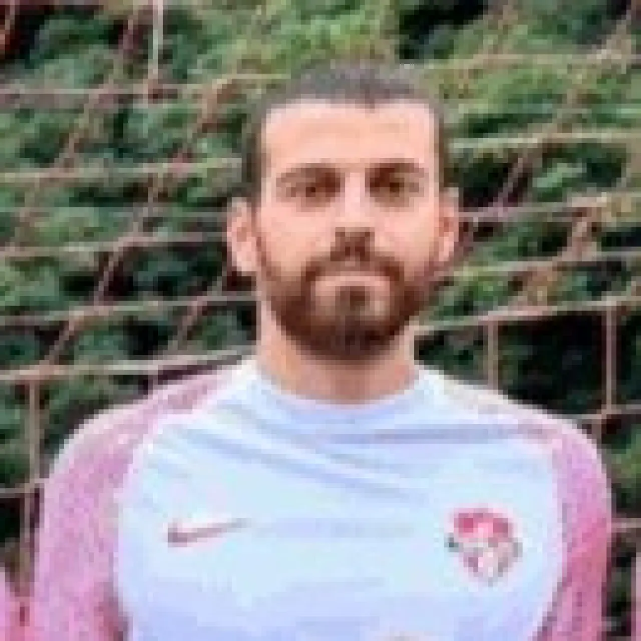 Porträt eines bärtigen Fußballspielers in weißem Trikot mit Vereinswappen vor einem Tornetz im Freien.