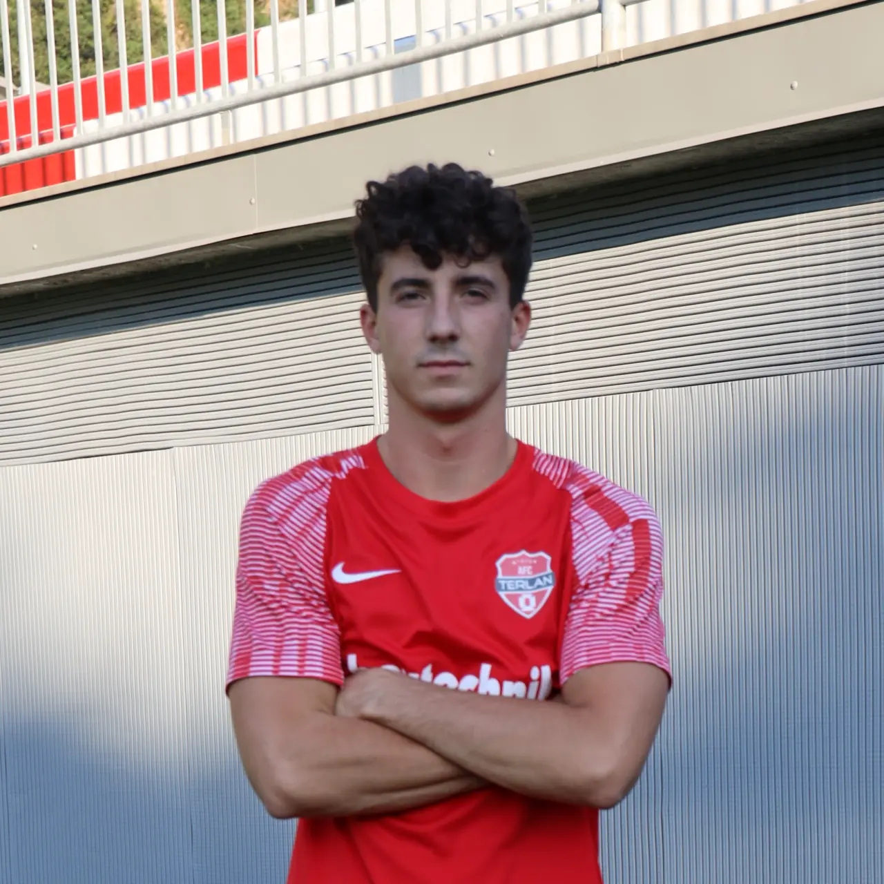 Ein junger Fußballspieler mit dunklen Locken steht mit verschränkten Armen vor einer grauen Wand im roten AFC-Terlan-Trikot mit Vereinswappen.