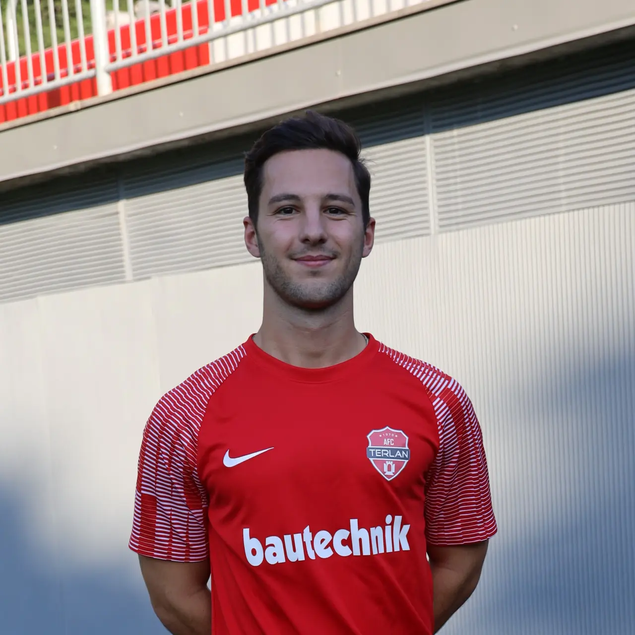 Porträt eines lächelnden Spielers des AFC Terlan im roten Trikot mit Vereinswappen und bautechnik-Sponsorenaufdruck vor einer hellen Wand.