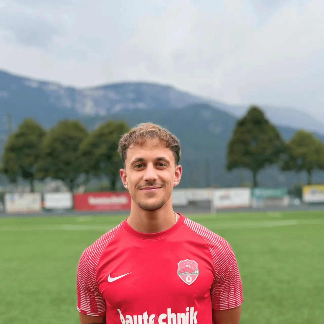 Porträt eines lächelnden Fußballspielers im roten AFC-Terlan-Trikot mit Bautechnik-Sponsor auf einem Kunstrasenplatz vor Bergen.