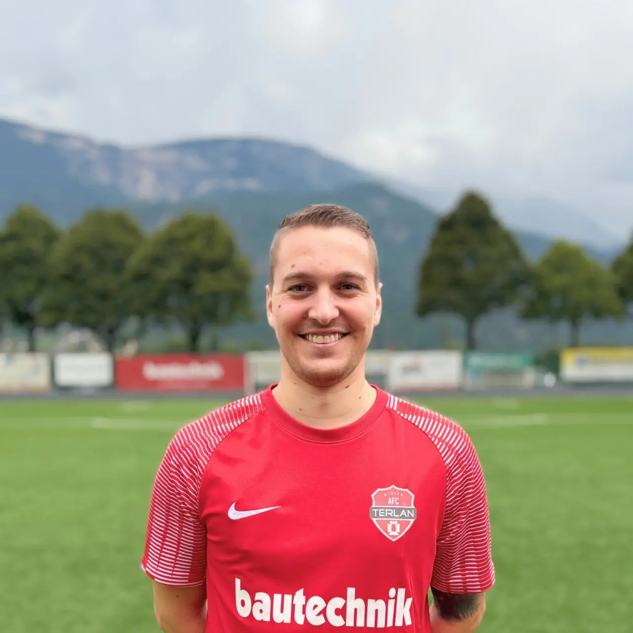 Lächelnder Spieler in roter Trikot des AFC Terlan mit Bautechnik-Sponsor auf einem Kunstrasenplatz vor Bergkulisse.