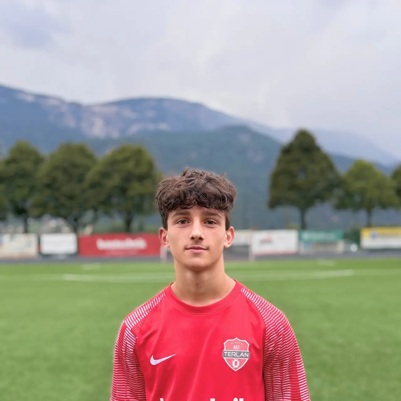 Porträt eines jungen Fußballspielers in roter Trikot mit Vereinswappen des AFC Terlan auf einem grünen Kunstrasenplatz vor Bergkulisse.