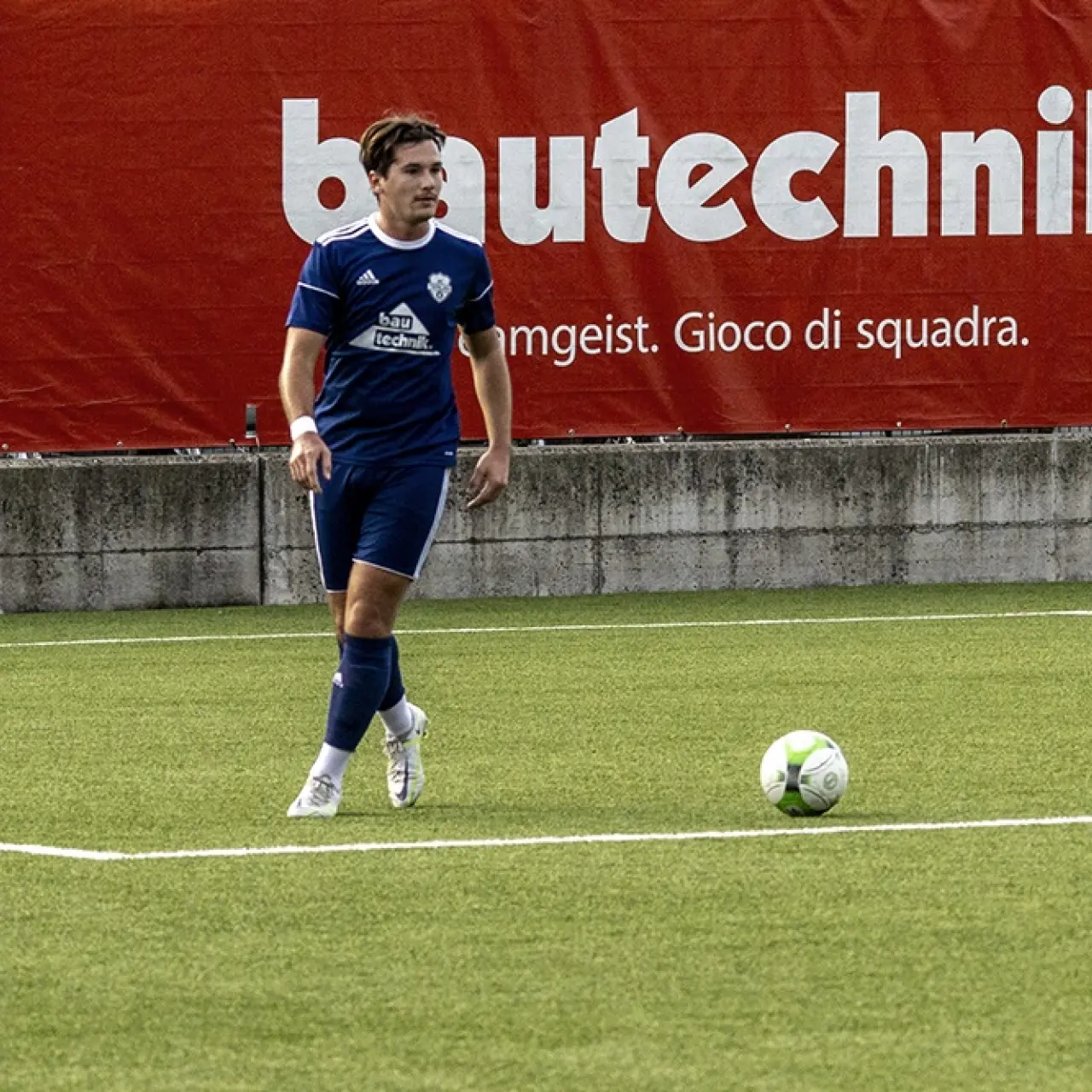 Spieler des AFC Terlan in blauem Trikot mit Bautechnik-Sponsorlogo führt den Ball auf dem Kunstrasen vor roter Werbebande.