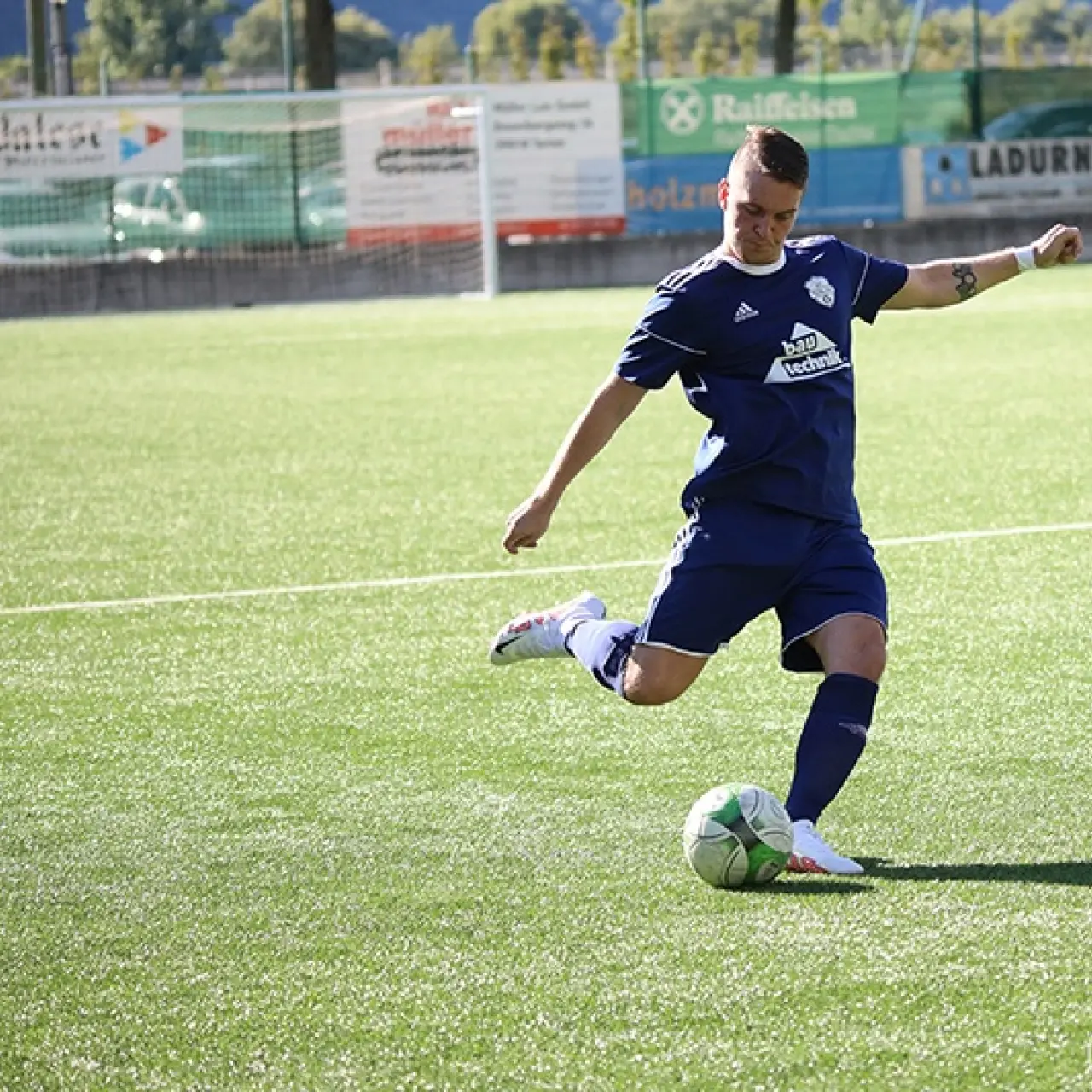 Spieler in dunkelblauem Trikot schießt auf dem Kunstrasen den Ball, im Hintergrund Bandenwerbung und ein Schiedsrichter.