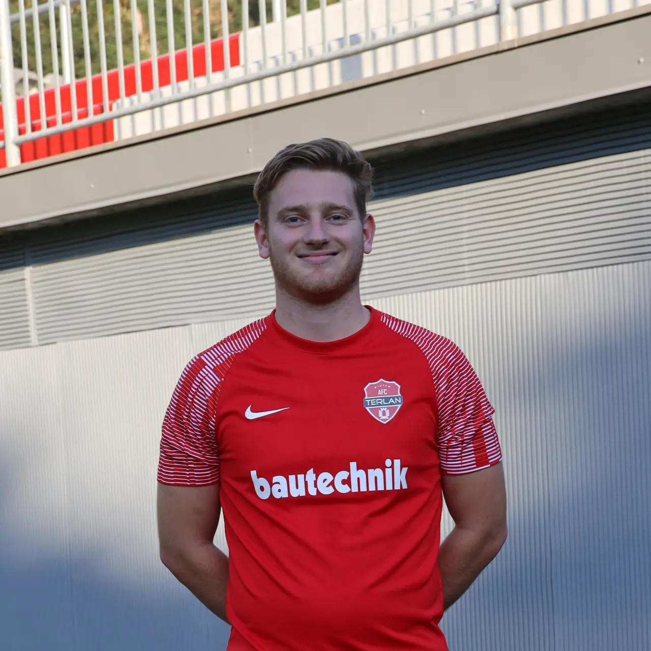 Spieler des AFC Terlan im roten Trikot mit Vereinswappen und Bautechnik-Sponsorenaufdruck, lächelnd vor einer grauen Wand.