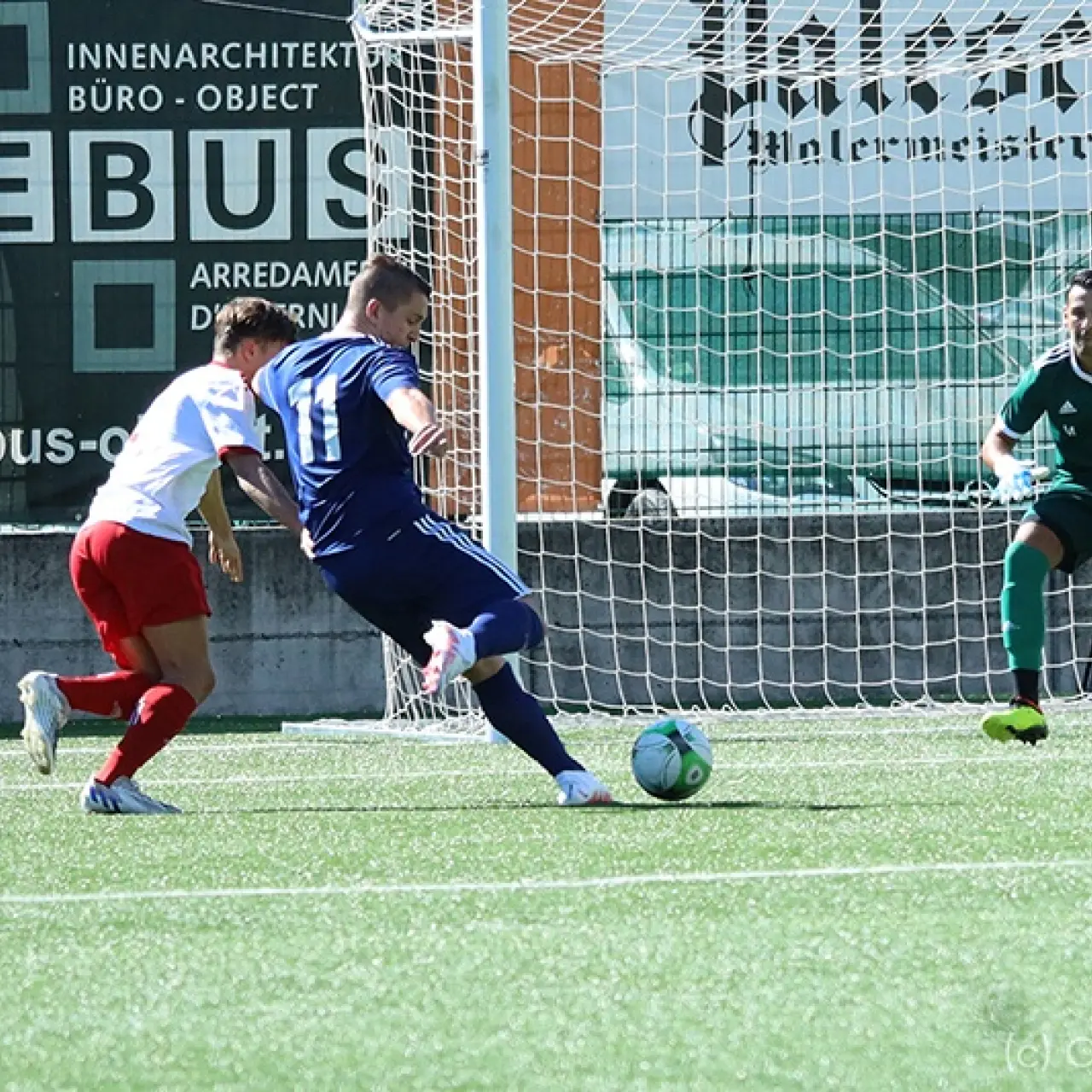 Spieler in blauem Trikot mit der Nummer 11 schießt auf das Tor, der grün gekleidete Torwart beobachtet die Szene, ein Gegenspieler in Rot-Weiß versucht zu stören.
