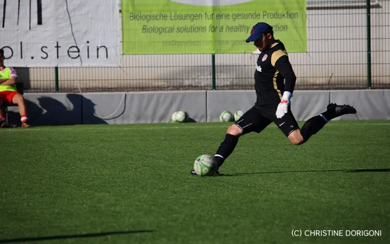 Torwart im schwarzen Trikot schießt auf dem Kunstrasen einen Ball ab, im Hintergrund Banner und Ersatzbälle am Spielfeldrand.