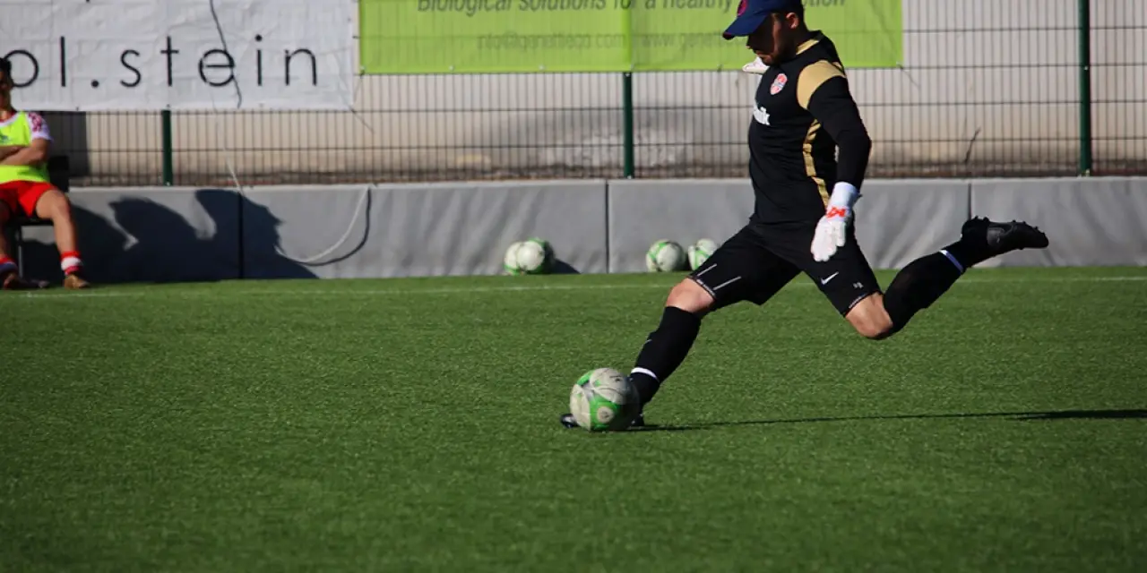 Torwart im schwarzen Trikot schießt auf dem Kunstrasen einen Ball ab, im Hintergrund Banner und Ersatzbälle am Spielfeldrand.