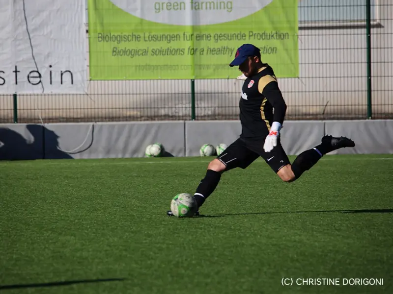 Torwart im schwarzen Trikot schießt auf dem Kunstrasen einen Ball ab, im Hintergrund Banner und Ersatzbälle am Spielfeldrand.