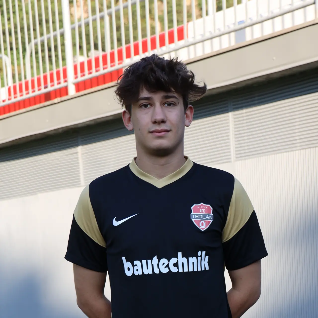 Porträt eines jungen Fußballspielers im schwarzen AFC-Terlan-Trikot mit goldenen Akzenten vor einer Stadiontribüne.