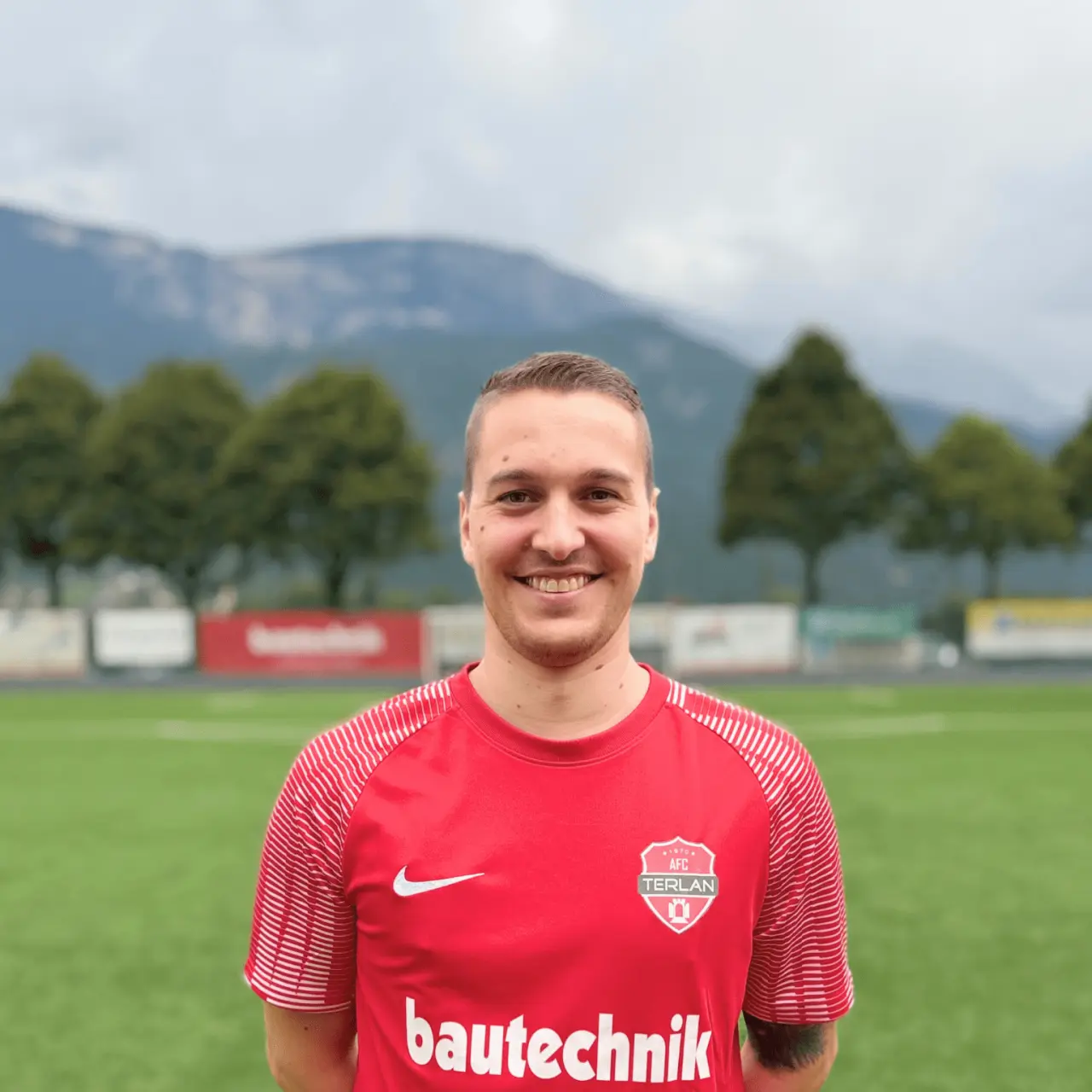 Lächelnder Spieler in roter AFC-Terlan-Trikot mit bautechnik-Sponsor vor einem Rasenplatz und Bergen im Hintergrund.