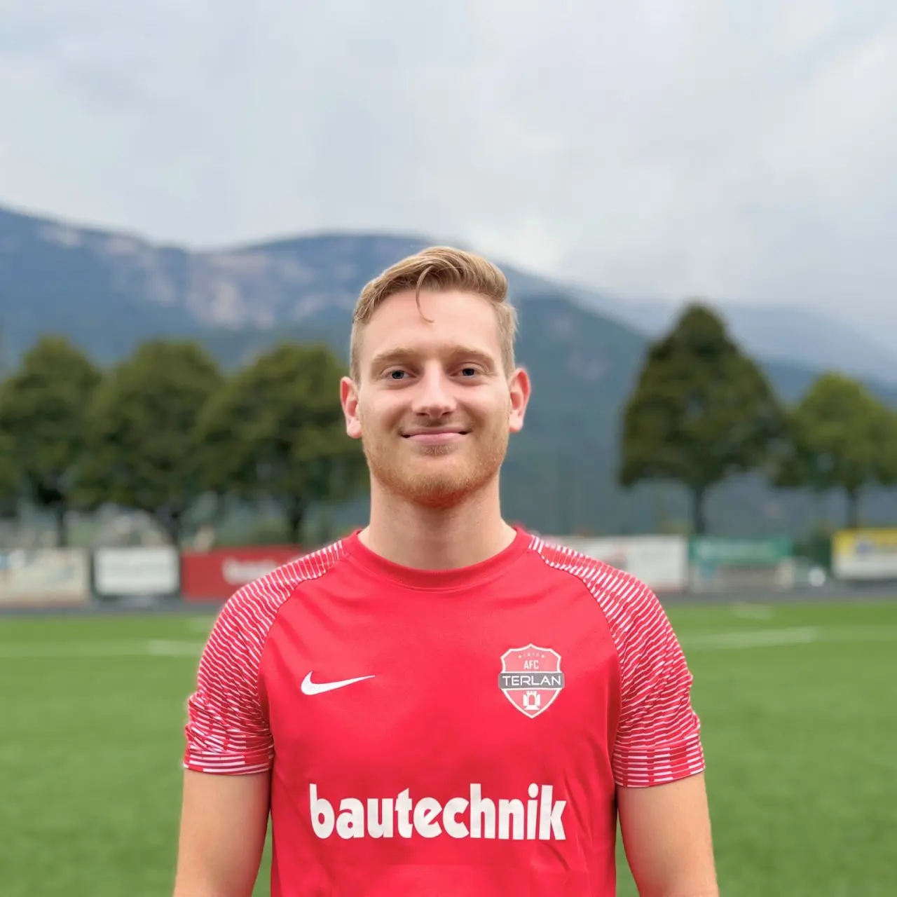 Porträt von Peter Gostner im roten Trikot des AFC Terlan mit Bautechnik-Sponsor auf einem Kunstrasenplatz vor Bergkulisse.