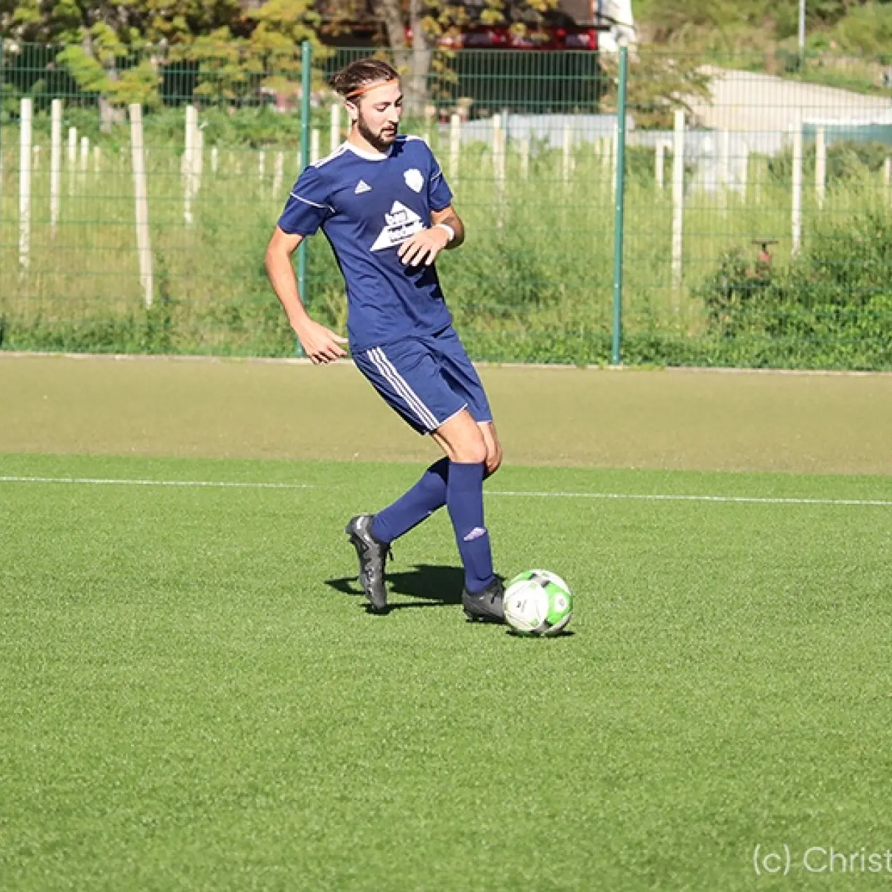 Ein Spieler in dunkelblauem Trikot führt auf dem grünen Kunstrasen den Ball mit dem Fuß am Platz entlang.