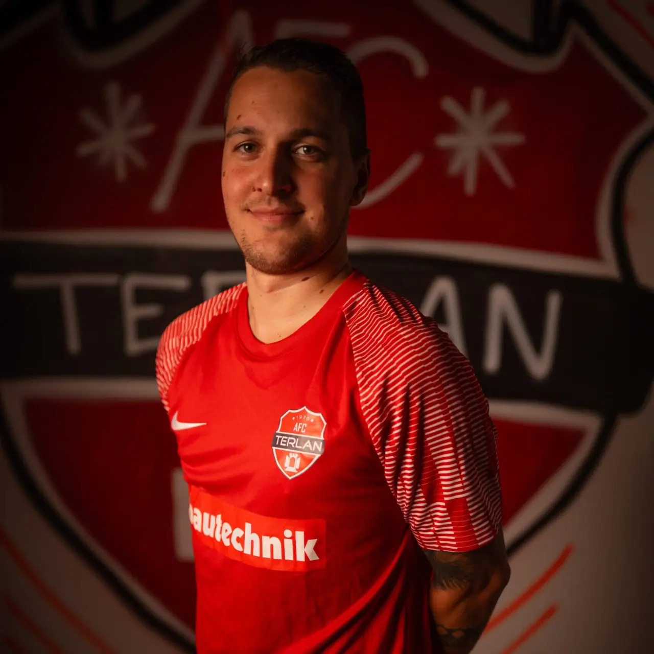 Porträt eines Spielers des AFC Terlan in roten Trikot mit Vereinswappen und Bautechnik-Sponsor vor dem Vereinslogo an der Wand.