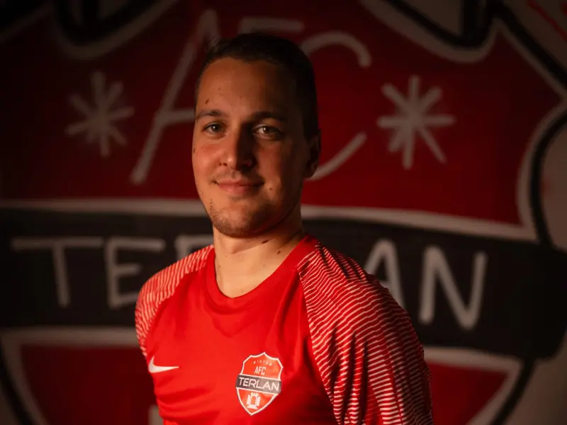 Porträt eines Spielers des AFC Terlan in roten Trikot mit Vereinswappen und Bautechnik-Sponsor vor dem Vereinslogo an der Wand.