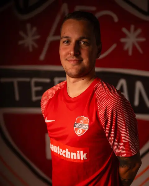 Porträt eines Spielers des AFC Terlan in roten Trikot mit Vereinswappen und Bautechnik-Sponsor vor dem Vereinslogo an der Wand.