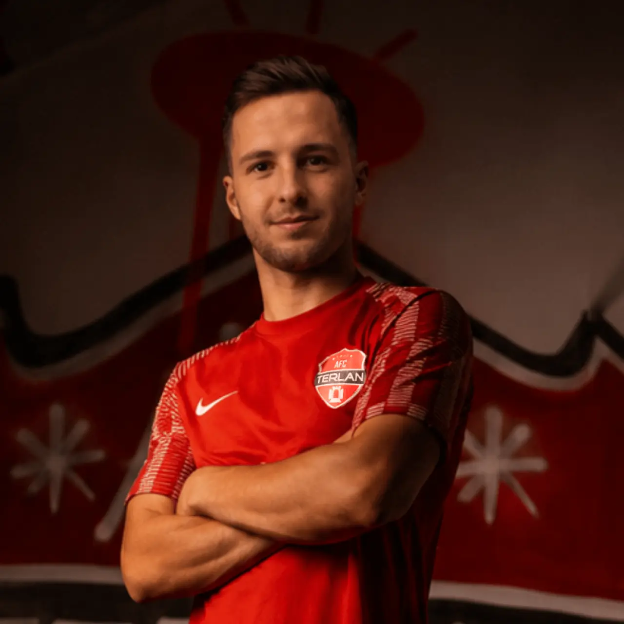 Spieler des AFC Terlan im roten Trikot mit Vereinswappen steht mit verschränkten Armen vor einer roten Wand mit Graffiti.