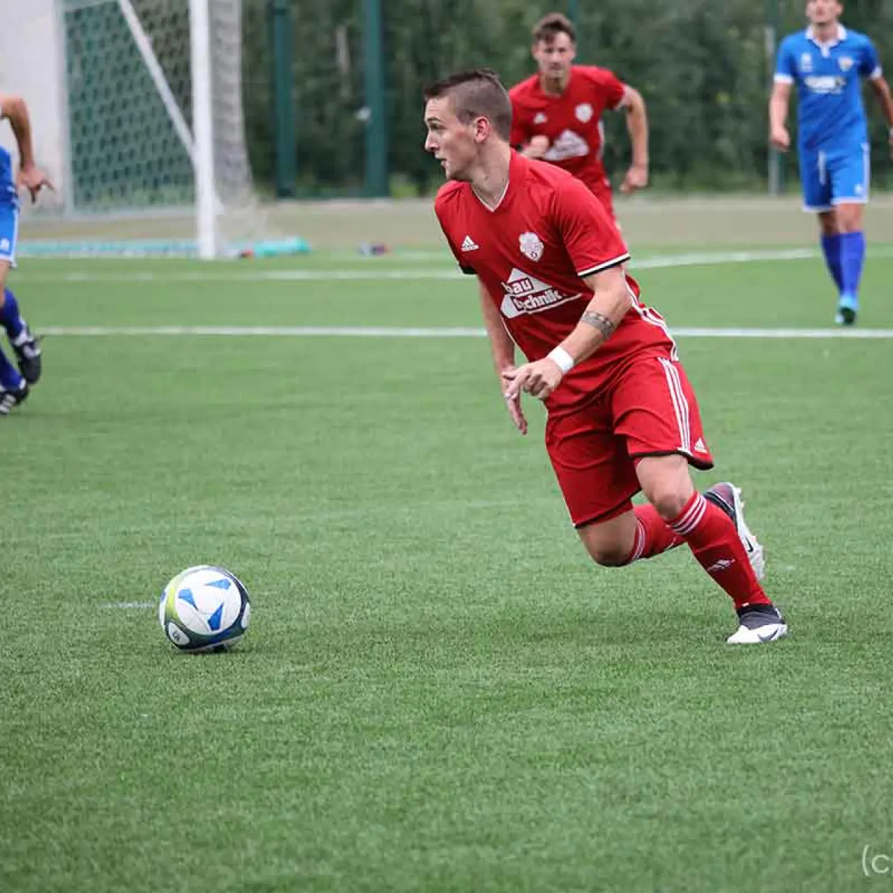Spieler des AFC Terlan in roter Trikot führt den Ball auf dem Kunstrasen, im Hintergrund Gegenspieler in Blau.