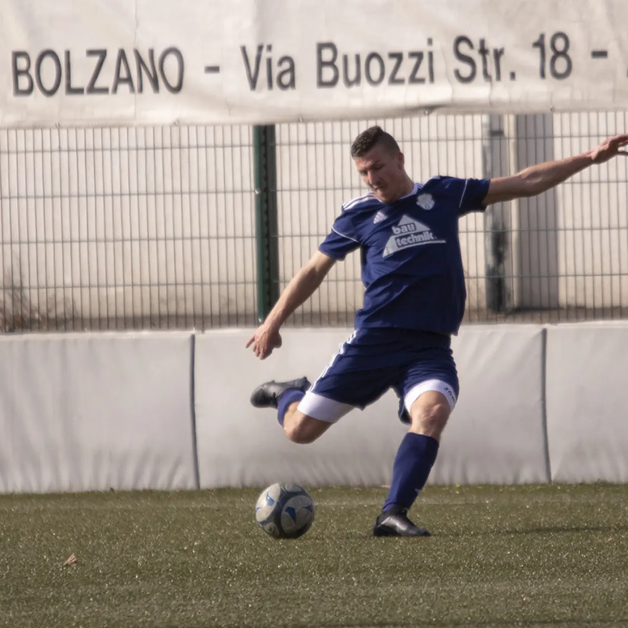Spieler in blauem Trikot mit Sponsorlogo Bautechnik schießt den Ball auf dem Kunstrasen, im Hintergrund Bande und Banner Bozen.