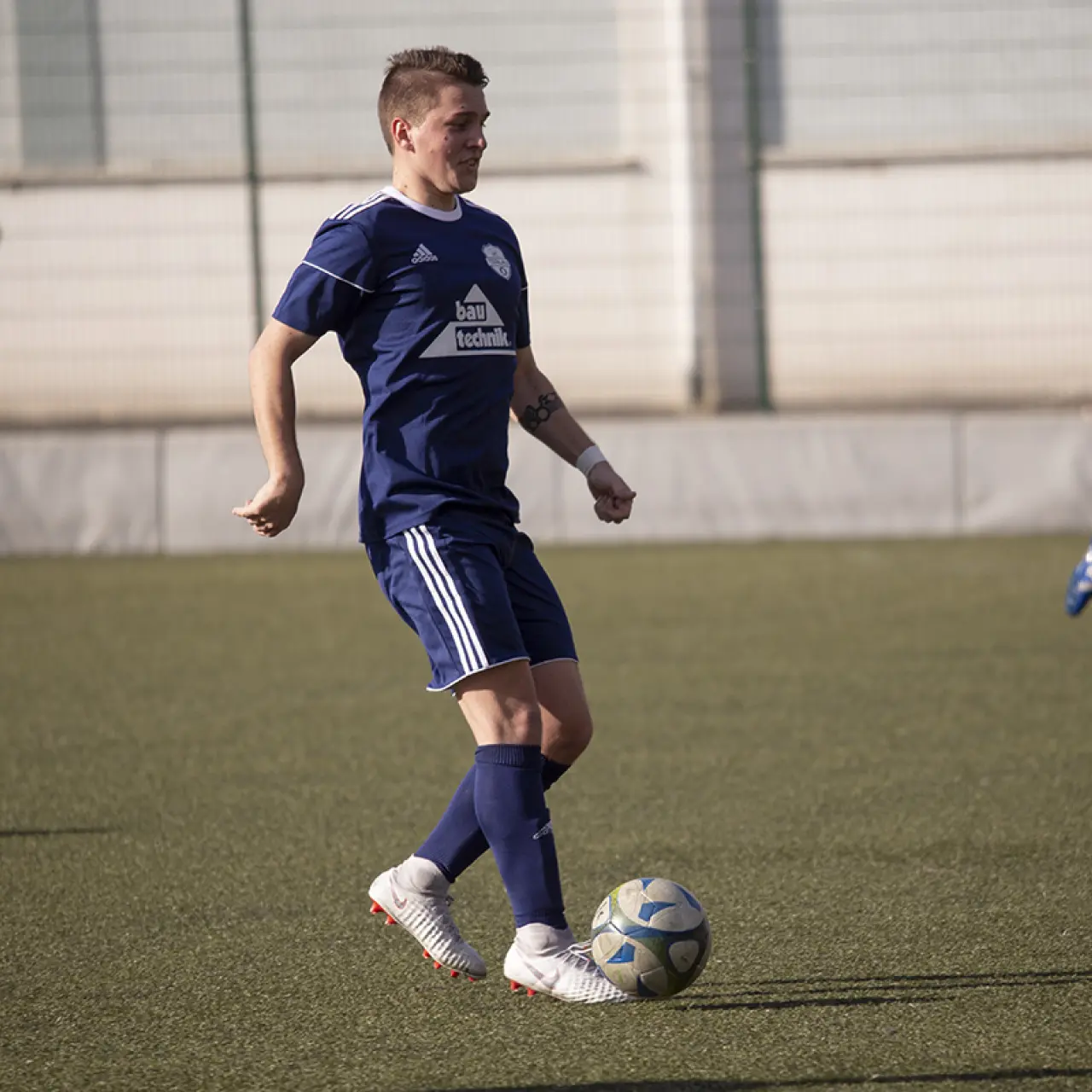 Spieler in blauem Trikot mit Bautechnik-Sponsorenlogo führt den Ball auf dem Rasen, im Hintergrund ein zweiter Spieler in Blau.
