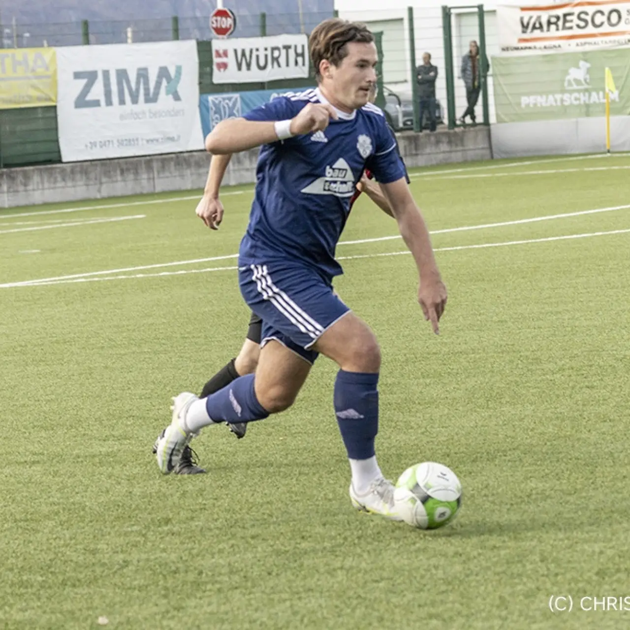 Spieler des AFC Terlan in blauem Trikot führt den Ball im Spiel gegen Partschins am Ballbesitz über den Rasen, Werbebanden im Hintergrund.