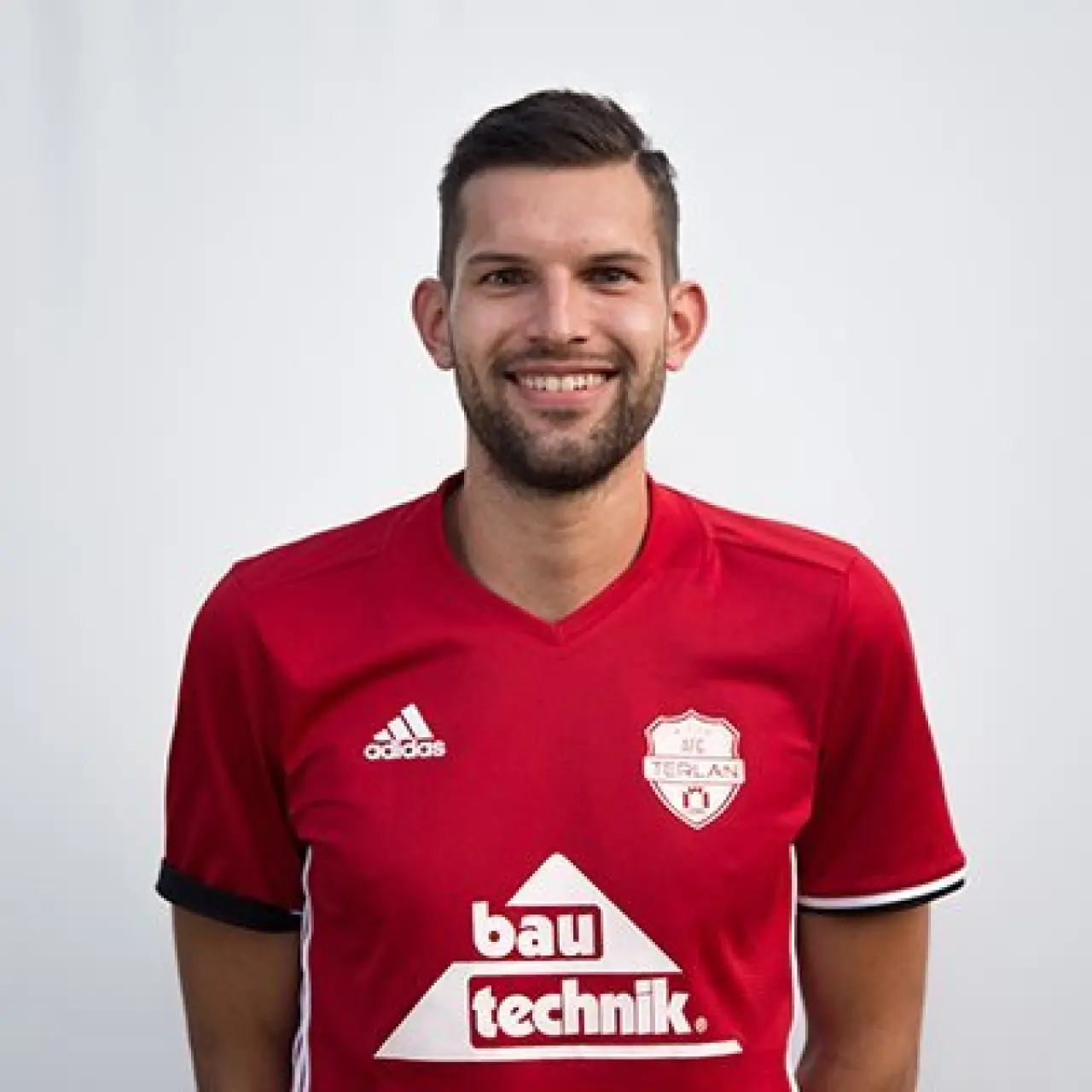 Porträt eines lächelnden Spielers des AFC Terlan im roten Trikot mit Vereinswappen und bautechnik-Sponsor vor hellem Hintergrund.
