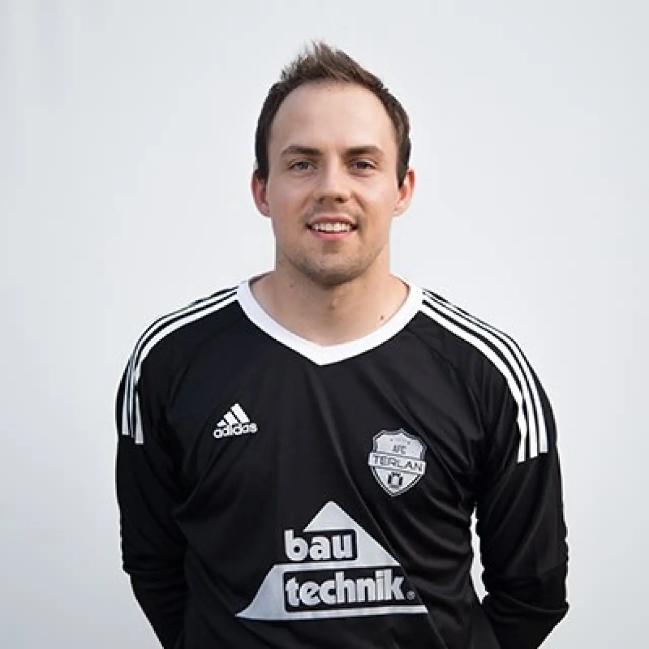 Porträt eines lächelnden Spielers in schwarzem Trikot mit Sponsorlogo bau technik und AFC-Terlan-Wappen vor hellem Hintergrund.