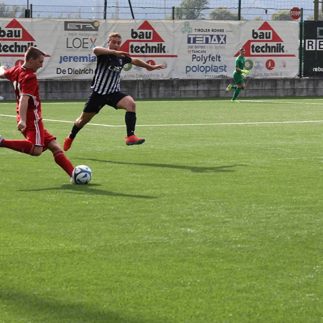Spielszene: Ein Spieler in roter Auswärtstrikot schießt den Ball, während ein Spieler in schwarz-weiß gestreiftem Trikot dagegen läuft, im Hintergrund Banden mit Sponsoren.