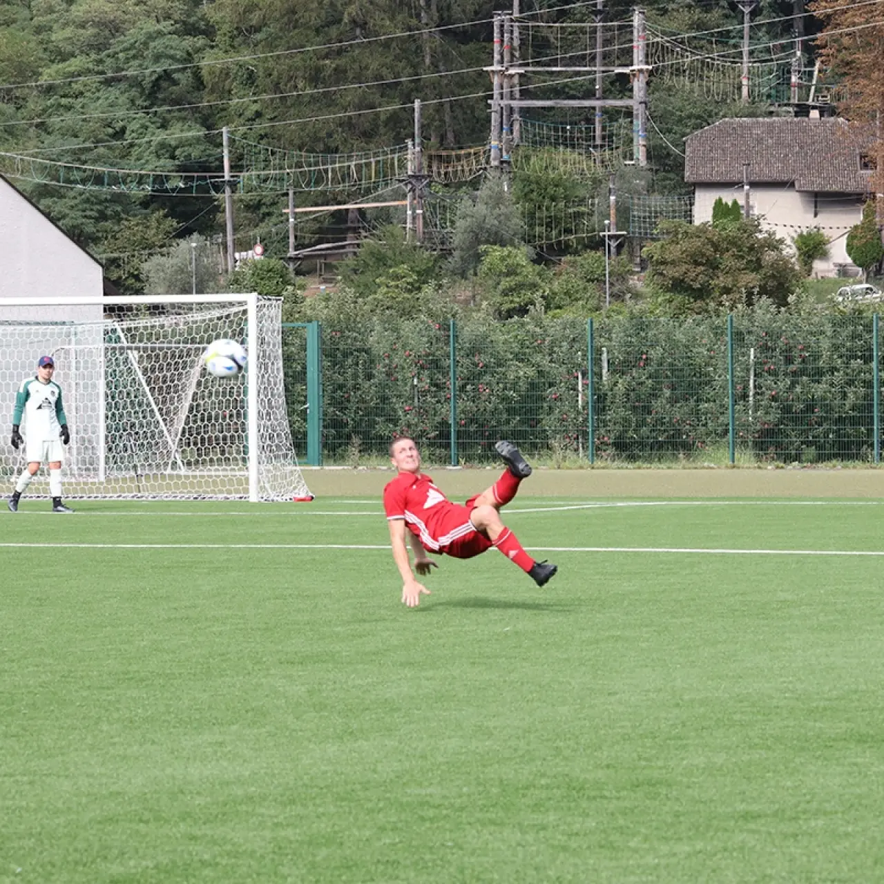 Ein Spieler in roter Trikot-Kombination streckt sich in der Luft zum Fallrückzieher, während der Torwart im Tor den Ball beobachtet.