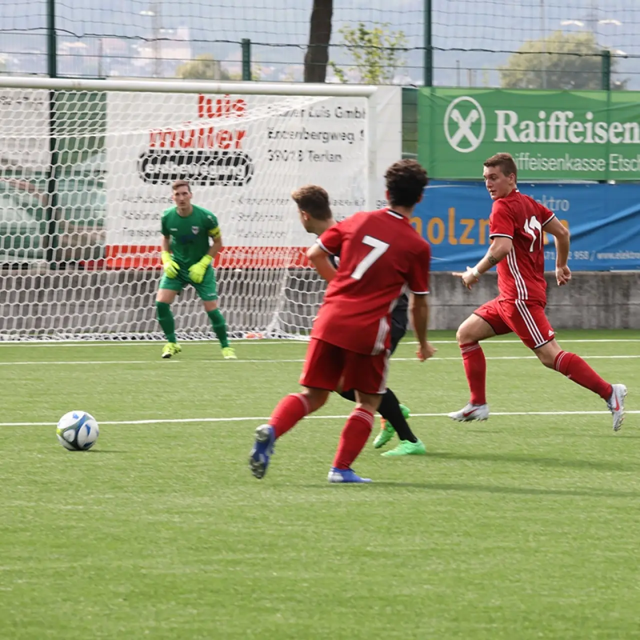 Spielszene auf dem Kunstrasenplatz: Zwei rote Spieler, darunter Nummer 7, laufen dem Ball nach, während der Torwart in Grün im Tor wartet.