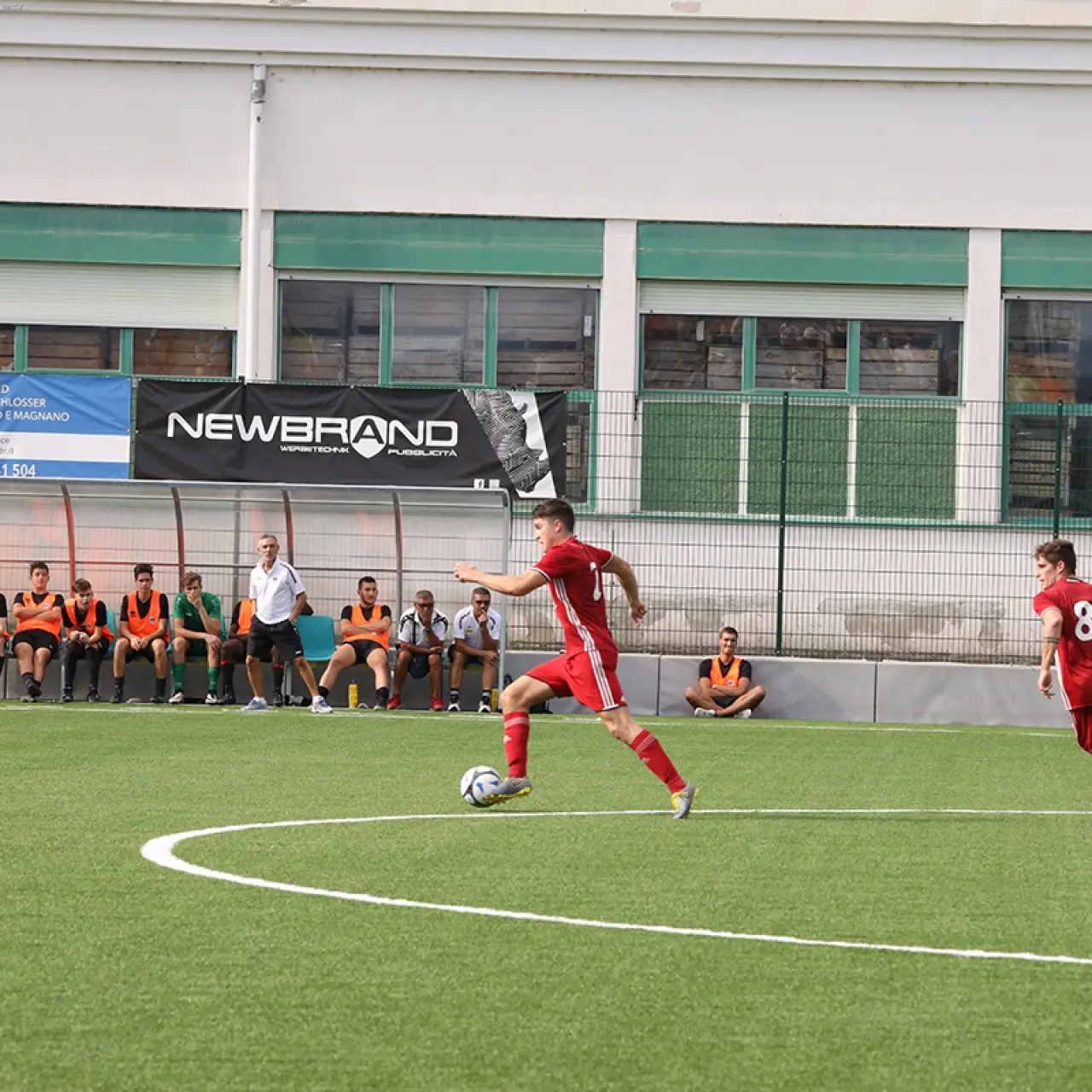Ein roter Spieler führt den Ball auf dem Kunstrasen, ein zweiter Spieler in Rot mit Nummer 8 läuft mit, im Hintergrund die Ersatzbank.