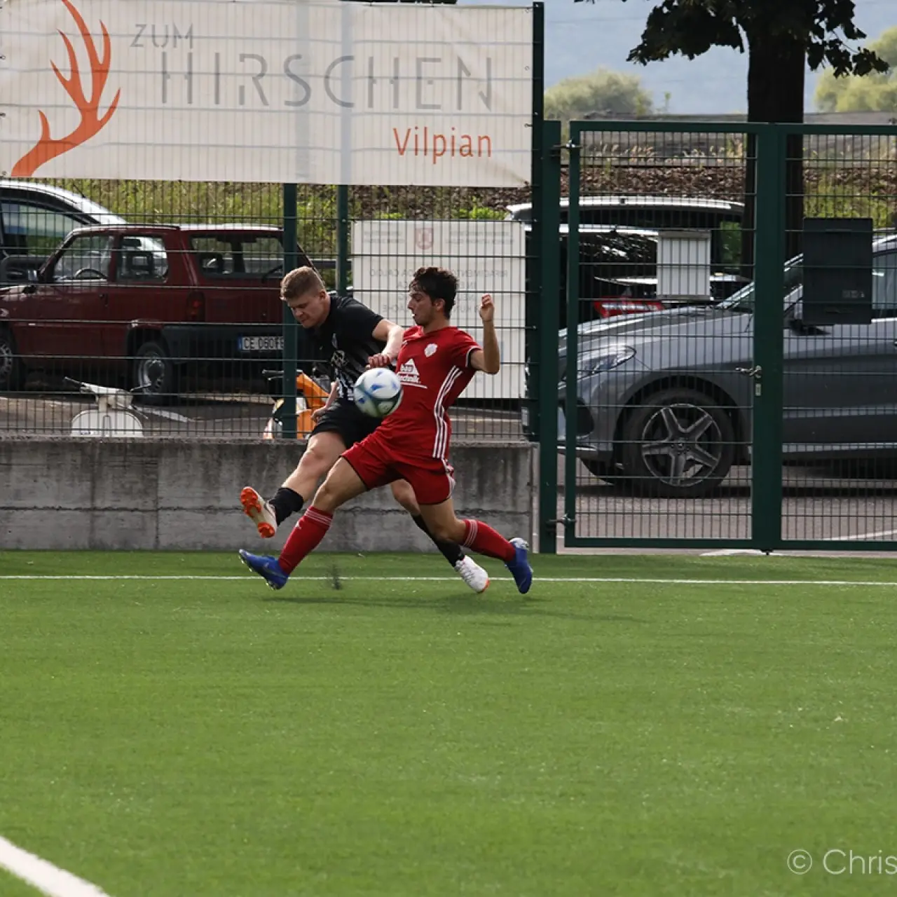 Zwei Fußballspieler kämpfen um den Ball auf einem Kunstrasenplatz, einer in Rot, einer in Schwarz, im Hintergrund ein Zaun und Werbebanner.