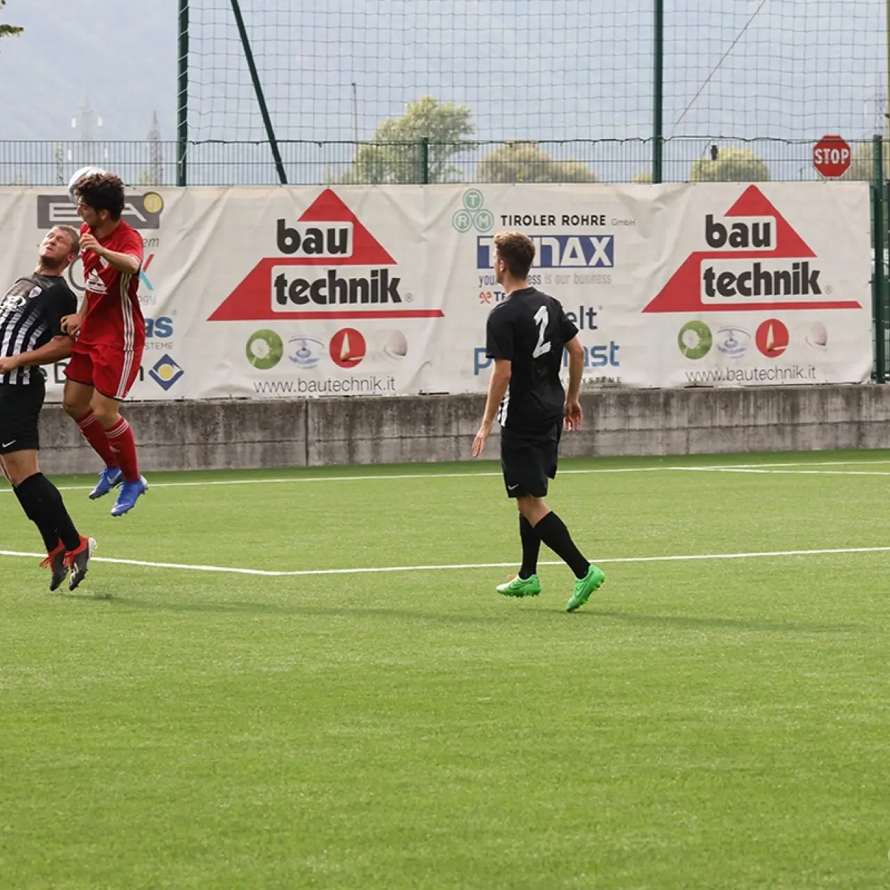 Zwei Spieler springen gemeinsam zum Kopfball, einer in Rot, einer in Schwarz-Weiß gestreift; im Hintergrund Bandenwerbung von Bautechnik und Rebus.