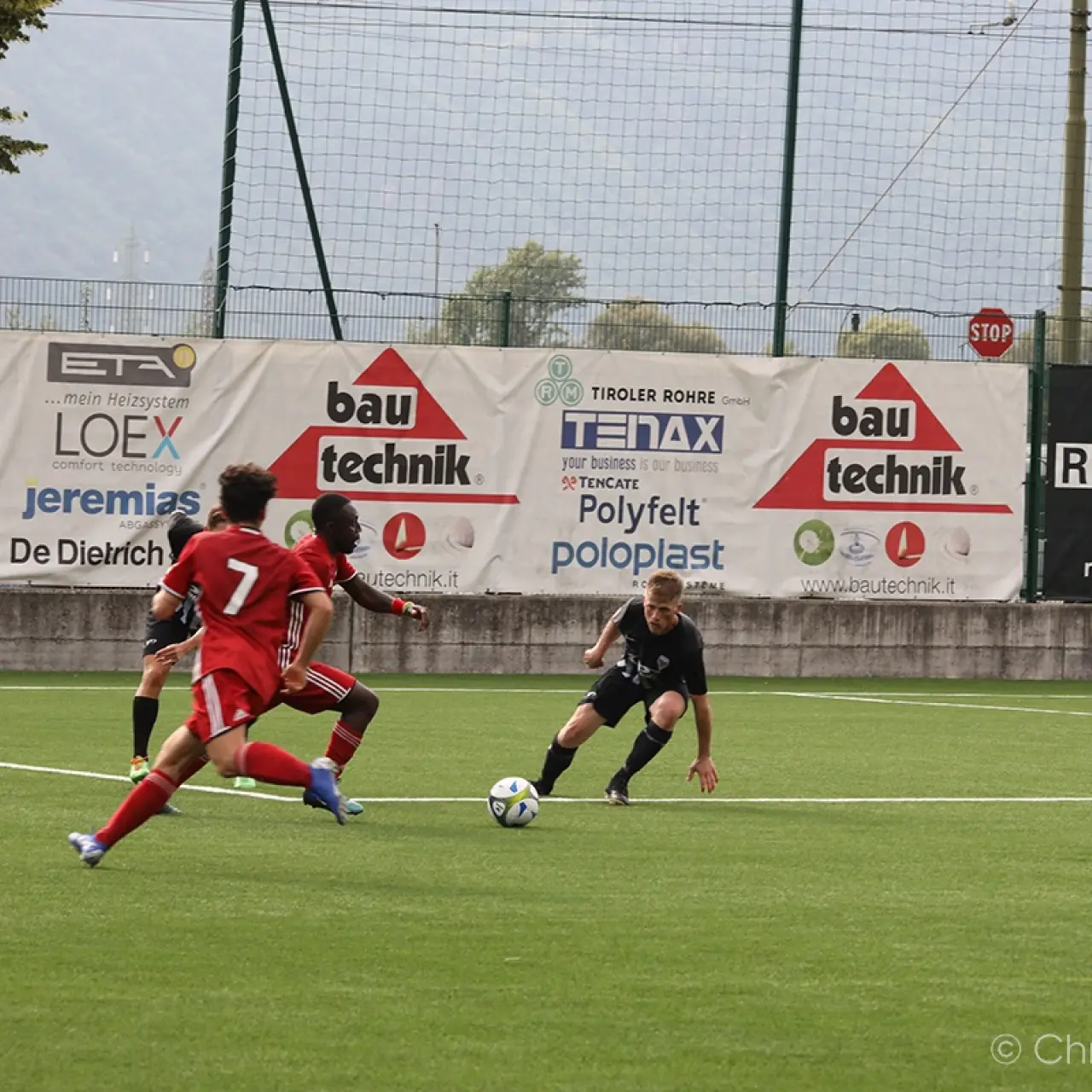 Spielszene: Ein Spieler in Schwarz führt den Ball am Boden, während zwei Spieler in Rot ihn bedrängen; im Hintergrund Werbebanden von bau technik.