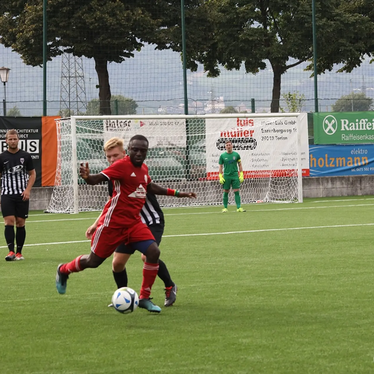 Spielszene: Ein Spieler in Rot führt den Ball vor einem Gegner in Schwarz-Weiß, im Hintergrund Banden der Sponsoren Raiffeisen und Ladurner.