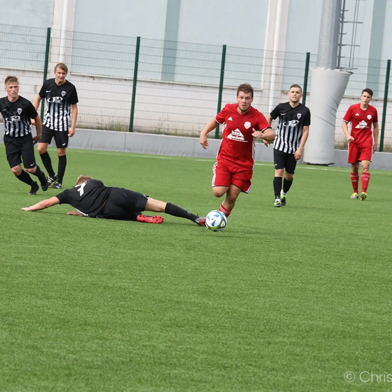 Spielszene: Ein roter Spieler führt den Ball am Gegner vorbei, während ein schwarz gekleideter Spieler am Boden rutscht und Gegenspieler mit gestreiften Trikots folgen.