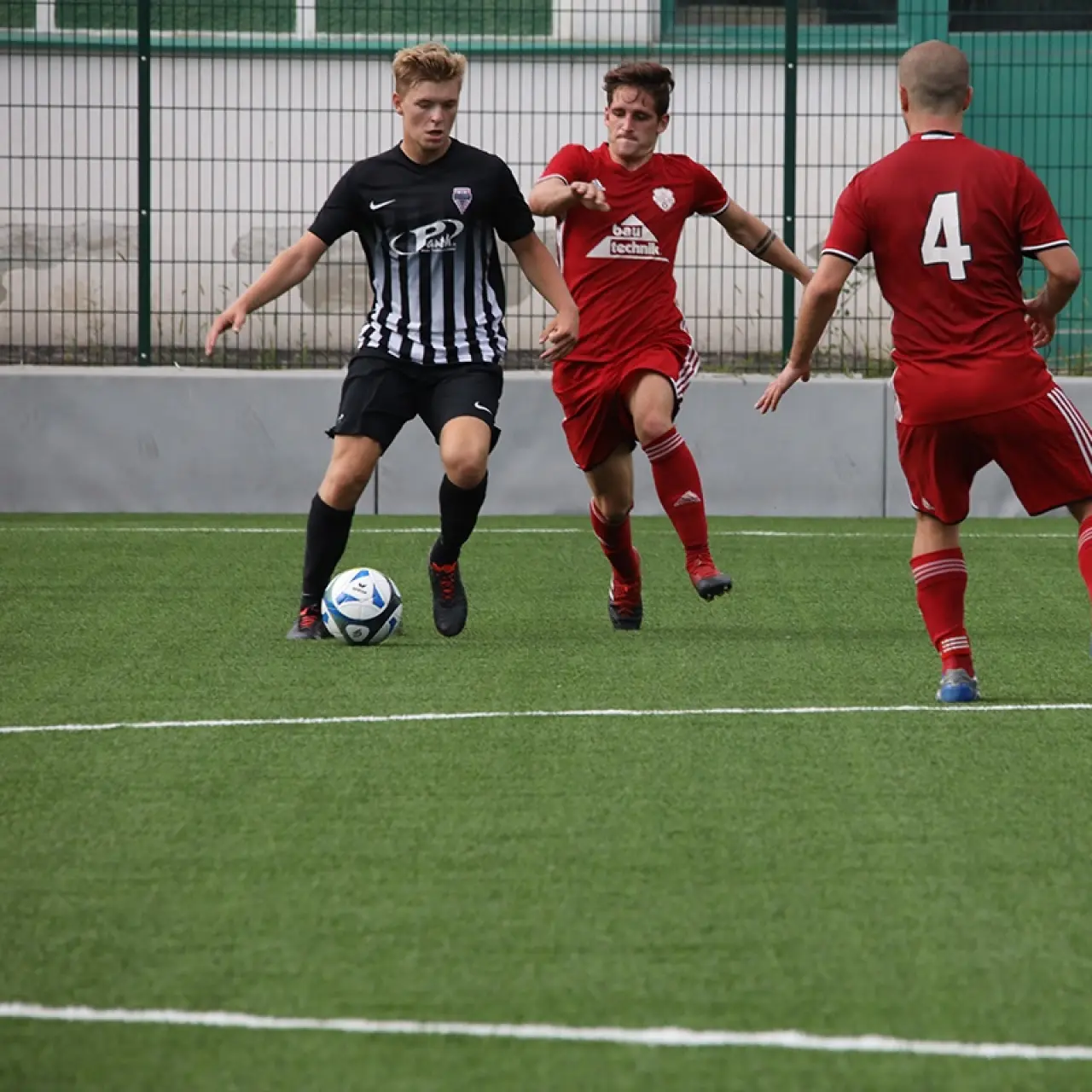 Spielszene auf dem Kunstrasenplatz: Ein Spieler in schwarz-weiß gestreiftem Trikot führt den Ball, während zwei Spieler in Rot ihn attackieren.