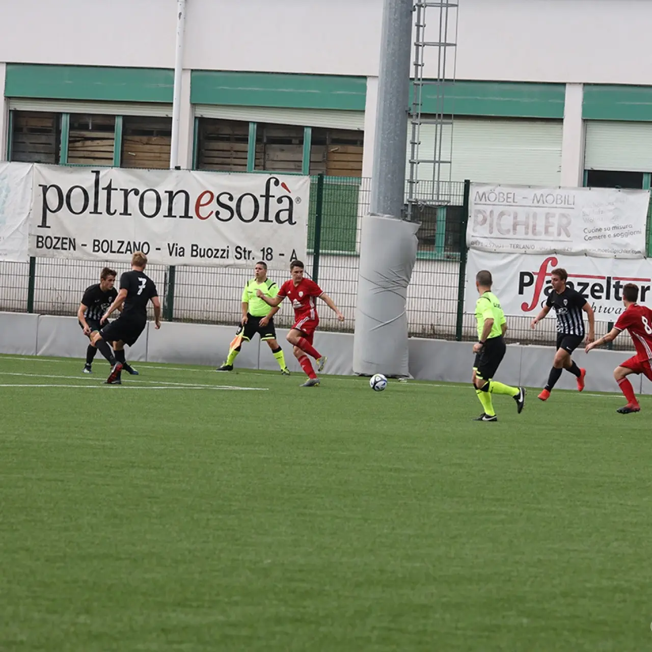 Spielszene auf dem Kunstrasen: Rot gekleidete Spieler des AFC Terlan verfolgen den Ball gegen Gegner in Schwarz-Weiß, Banden mit Sponsorenwerbung im Hintergrund.