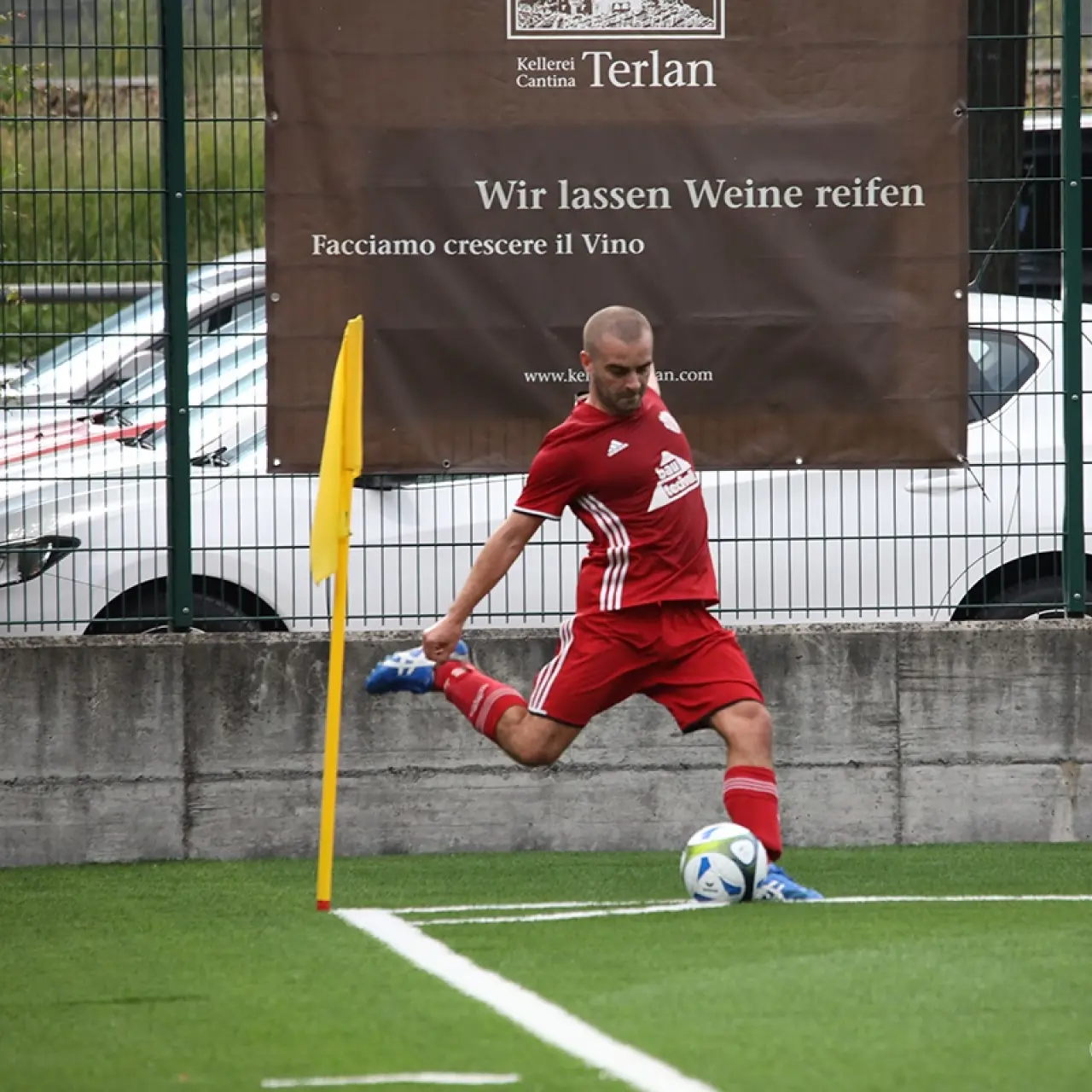 Spieler in roter Vereinskleidung schießt einen Eckball auf dem Kunstrasen, im Hintergrund Bande mit Werbung der Kellerei Terlan.