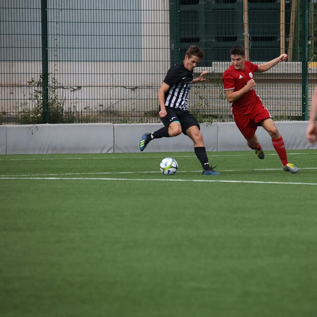 Spielszene: Ein schwarz-weiß gestreifter Spieler führt den Ball am Gegner in Rot vorbei, im Vordergrund ein Spieler mit Trikotnummer 6.