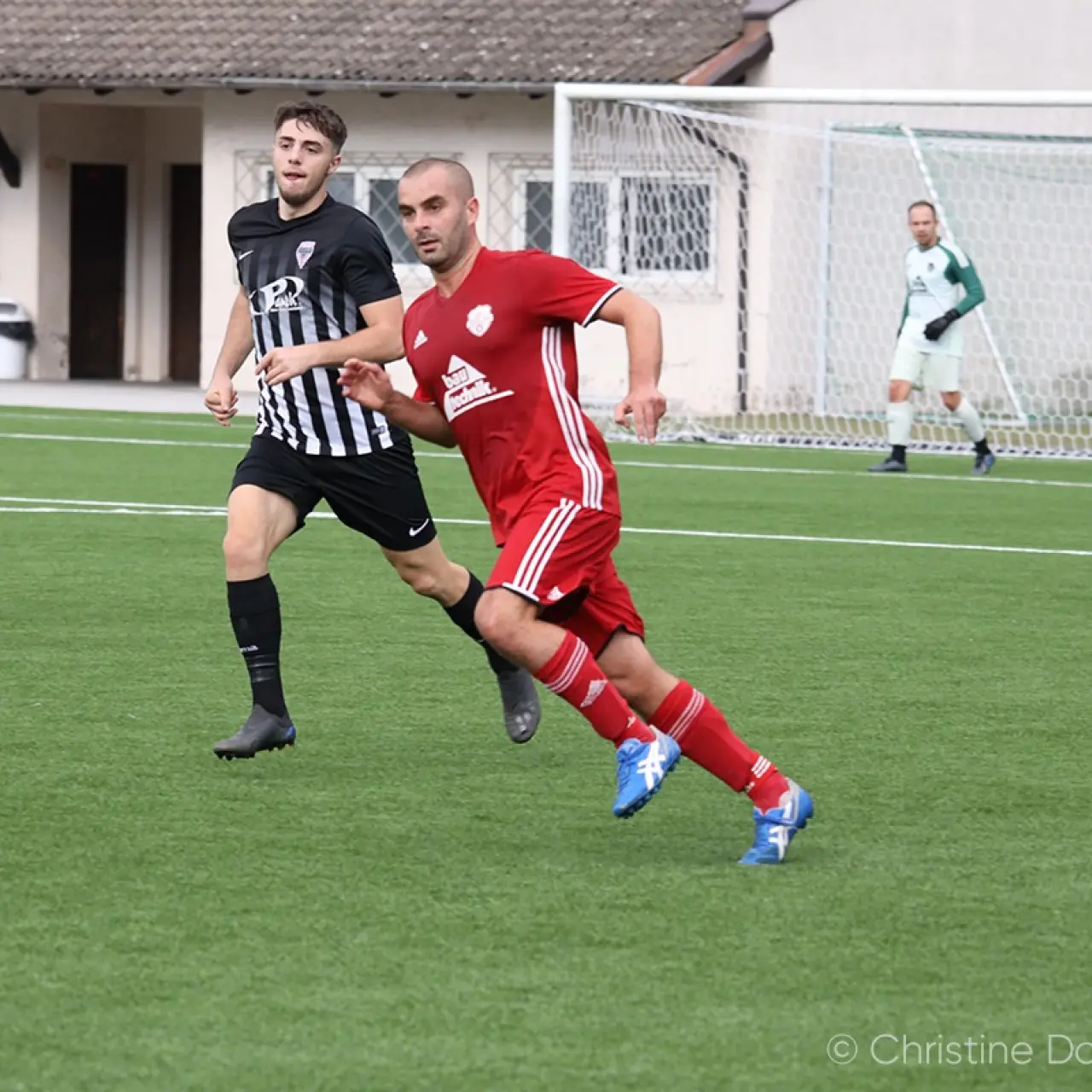 Spielszene: Ein roter Spieler des AFC Terlan läuft mit dem Ball am Fuß an einem Gegner in schwarz-weiß gestreiftem Trikot vorbei, der Torhüter wartet im Hintergrund.