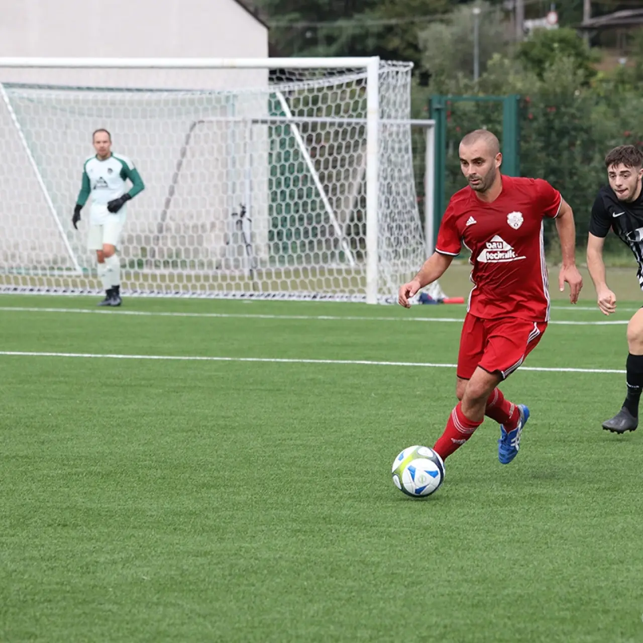 Ein Spieler in roter Trikot führt den Ball auf dem Kunstrasen, während ein Gegenspieler in Schwarz-Weiß folgt; im Hintergrund steht ein Torwart im Tor.