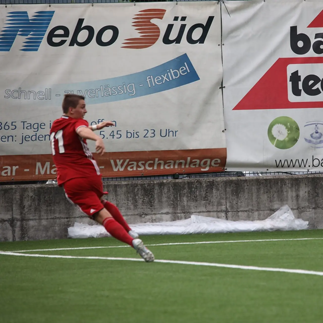 Spieler in roter Vereinskleidung mit Nummer 11 läuft auf dem Kunstrasen an einer Bande mit Werbebannern von Bautechnik und Nebo Süd vorbei.