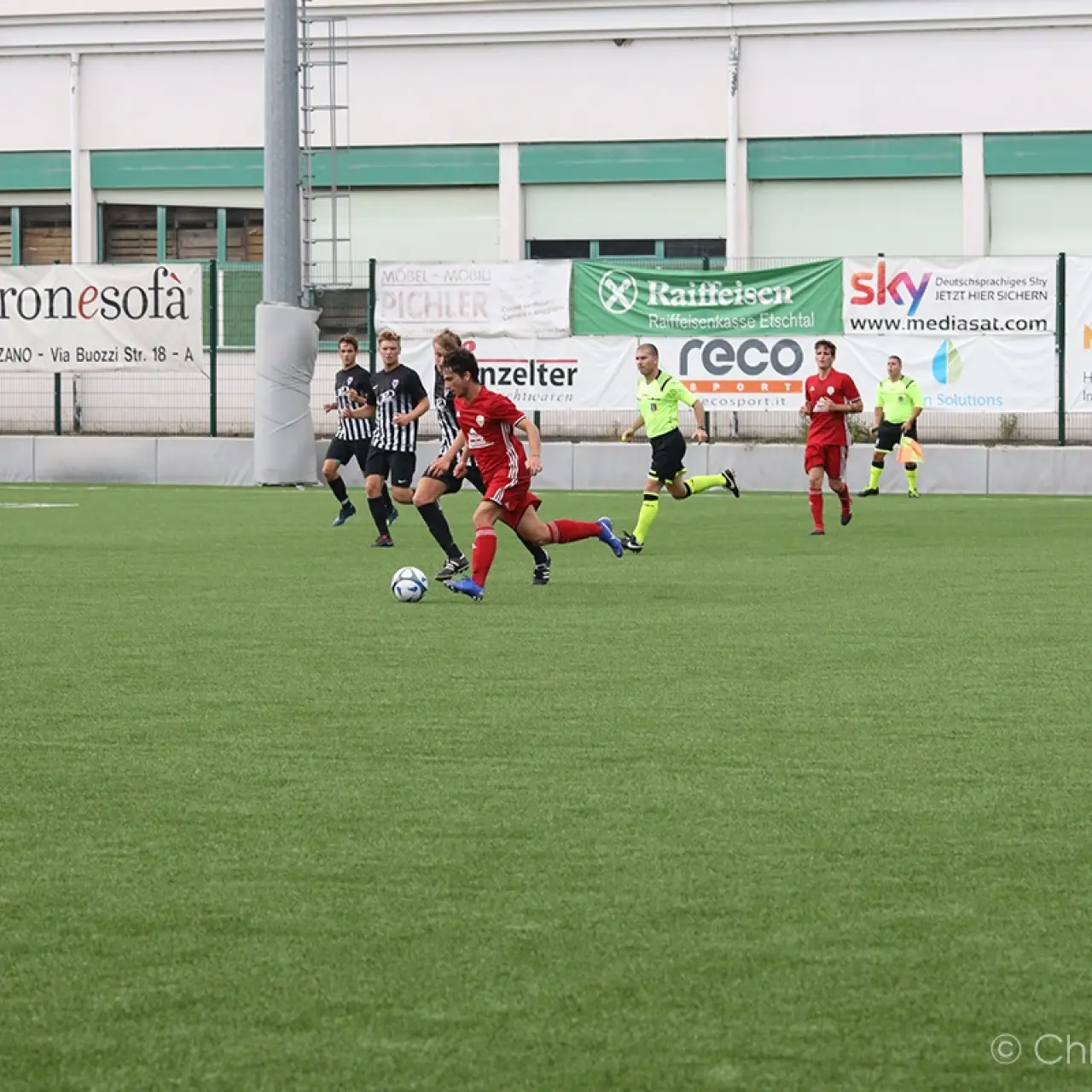 Spielszene auf dem Kunstrasen: Ein roter Spieler führt den Ball gegen zwei schwarz-weiße Gegenspieler, im Hintergrund Bandenwerbung und ein Schiedsrichter in Neongelb.