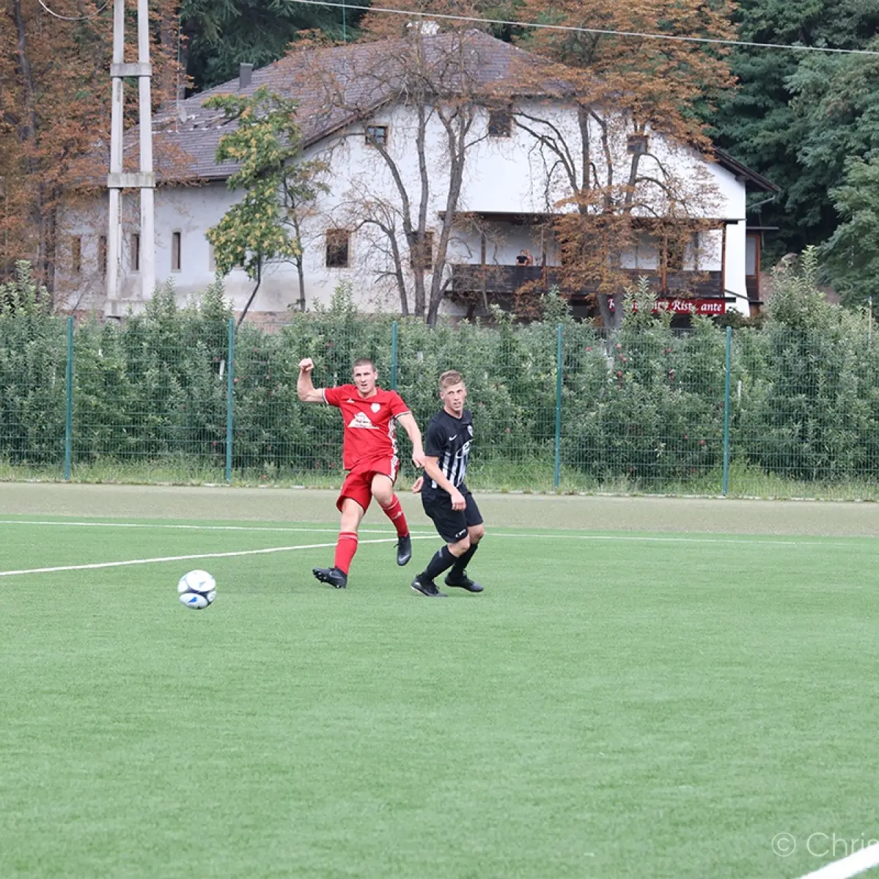 Zwei Fußballspieler im Zweikampf auf dem Kunstrasenplatz: links ein Spieler in Rot, rechts einer in Schwarz-Weiß gestreift, der Ball rollt vor ihnen.