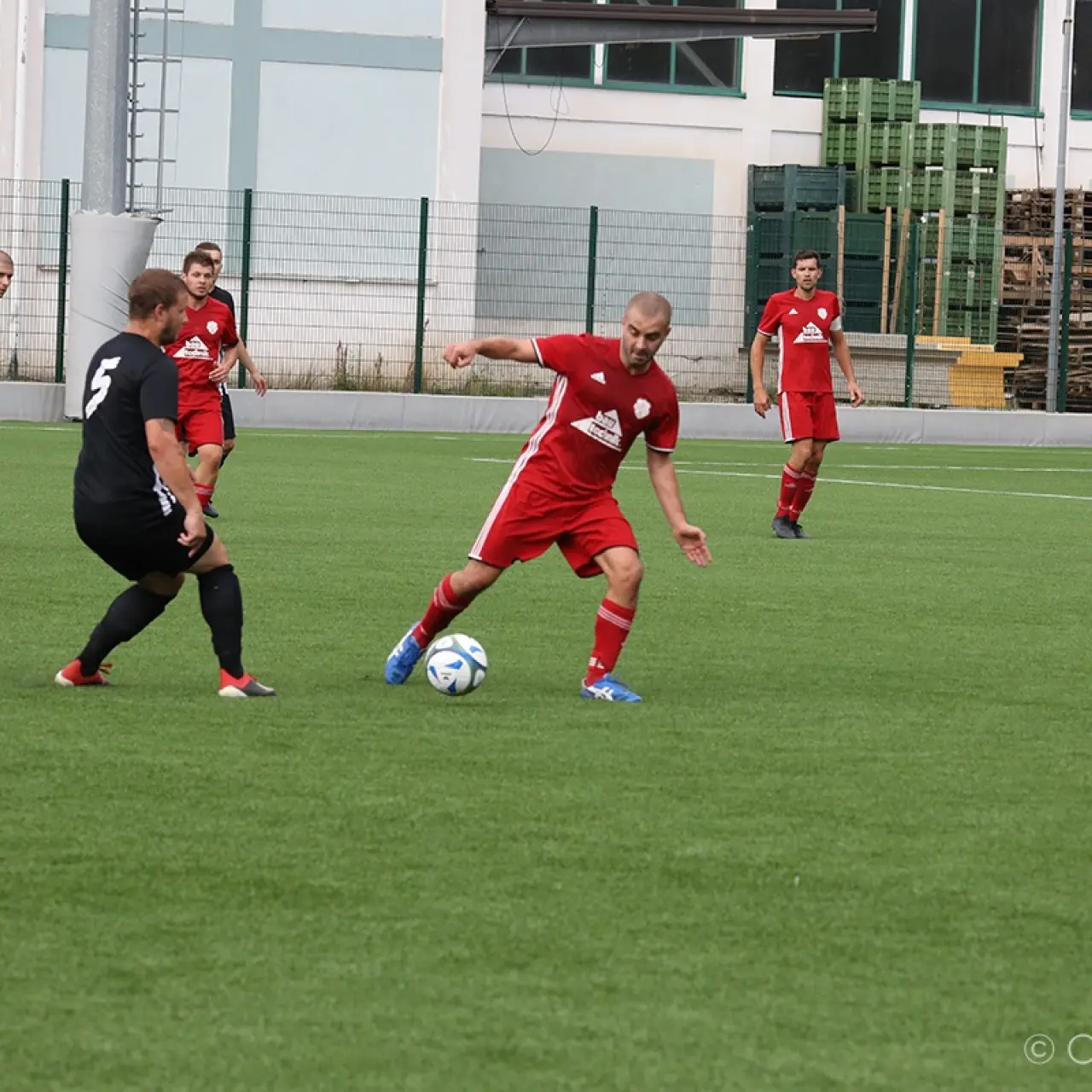 Spielszene: Ein Spieler in Rot führt den Ball am Boden, während ein Gegenspieler in Schwarz auf dem Kunstrasen auf ihn zuläuft.