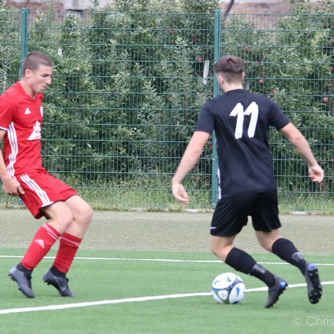 Zwei Spieler im Zweikampf auf dem Kunstrasen: Ein Spieler in Rot verfolgt den Ball, der Spieler in Schwarz mit der Nummer 11 führt ihn am Fuß.