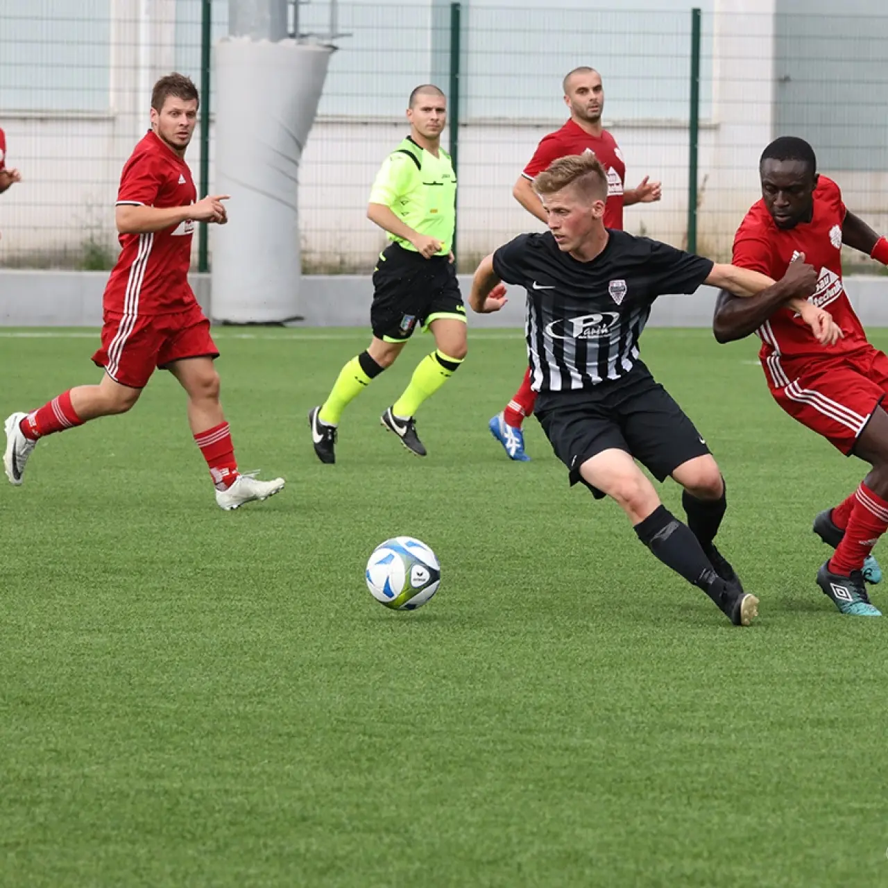 Spielszene: Ein schwarz-weiß gestreifter Spieler behauptet den Ball gegen einen Gegner in Rot auf einem Kunstrasenplatz, Schiedsrichter im Hintergrund.